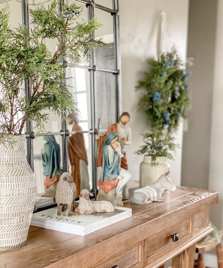 A foto mostra um presépio delicado sobre um aparador de madeira. As figuras de Maria, José e o Menino Jesus são acompanhadas por ovelhas e um burro. Um espelho reflete a cena, ao lado de um vaso com folhagens. O ambiente é decorado de forma acolhedora e serena.