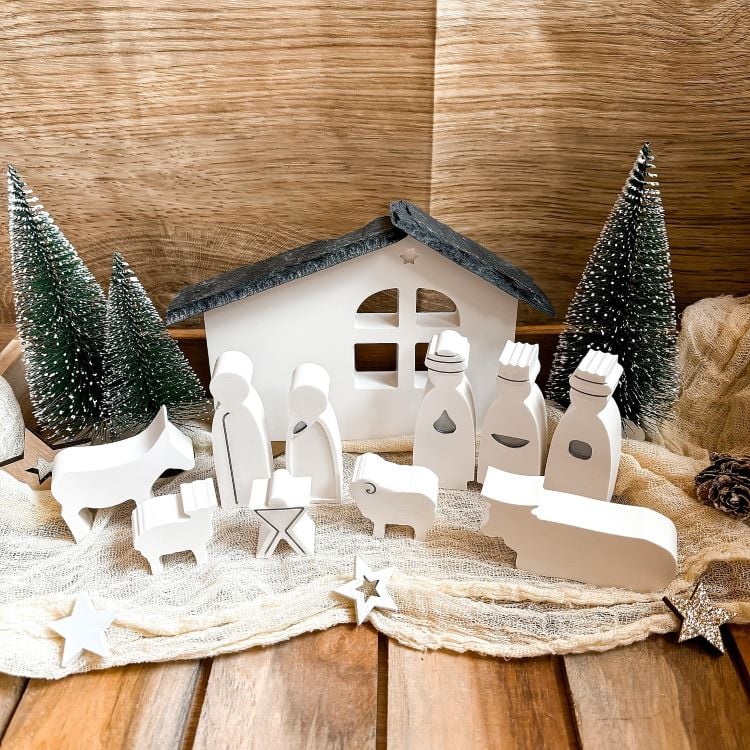 A imagem retrata um presépio minimalista com figuras brancas de madeira, incluindo a Sagrada Família, Reis Magos e animais. Ao fundo, uma cabana estilizada e pequenas árvores de Natal completam a composição. A cena está disposta sobre uma superfície de madeira, decorada com estrelas e tecido claro.