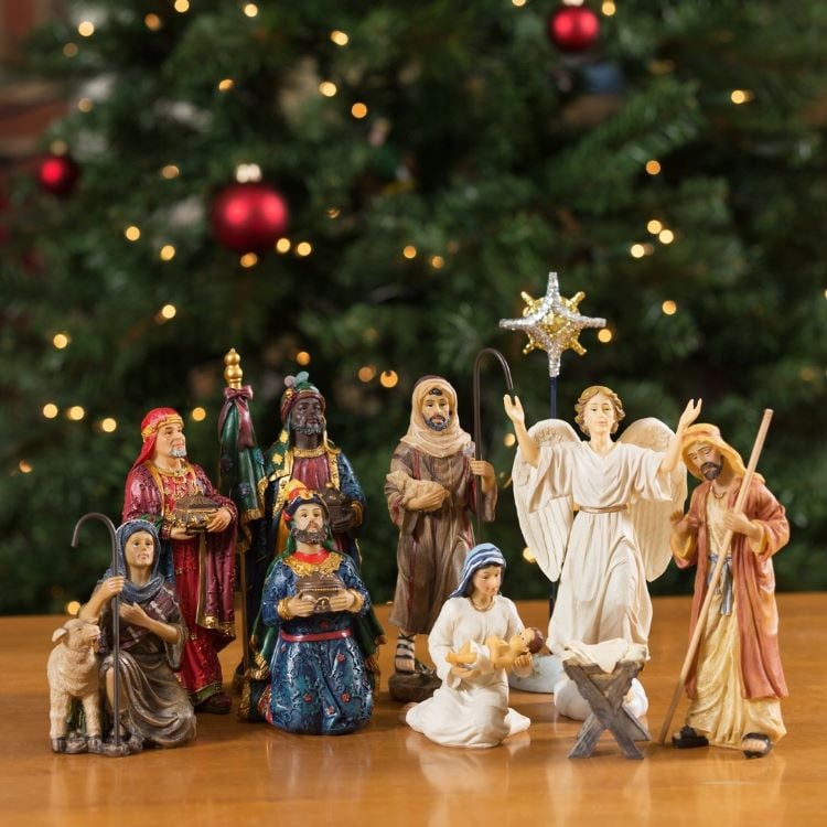 A foto apresenta um presépio com detalhadas figuras coloridas. Estão presentes Maria, José, o Menino Jesus, os Três Reis Magos, um anjo e pastores. Ao fundo, uma árvore de Natal decorada com luzes e bolas vermelhas adiciona um toque festivo ao ambiente, criando uma cena acolhedora.