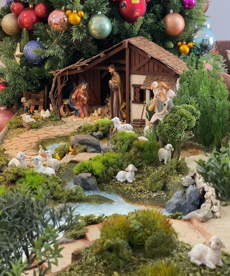 A imagem mostra um presépio detalhado sob uma árvore de Natal decorada com bolas coloridas. A cena inclui a Sagrada Família, cercada por ovelhas e árvores em miniatura. Um pequeno riacho com patos e patinhos atravessa o cenário, criando uma atmosfera de tranquilidade e beleza natural.