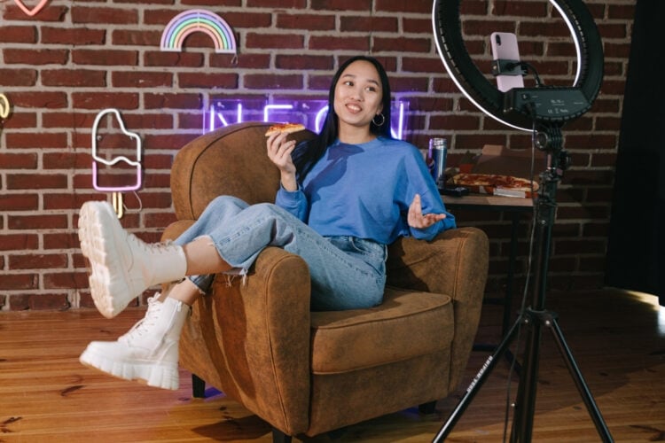 A imagem mostra uma mulher sentada em uma poltrona, comendo pizza, enquanto fala para uma câmera em um tripé com ring light. O ambiente tem parede de tijolos com luzes de neon coloridas, criando uma atmosfera descontraída e moderna.