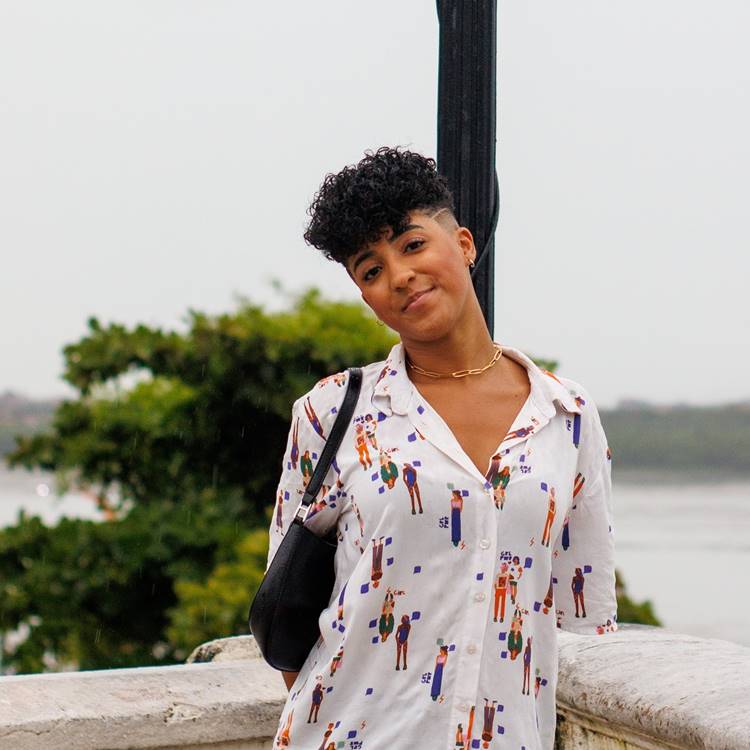 A imagem mostra uma pessoa sorrindo, com cabelo curto e encaracolado. Ela usa uma camisa estampada com figuras coloridas e carrega uma bolsa preta. O fundo é ao ar livre, com árvores e água ao longe.