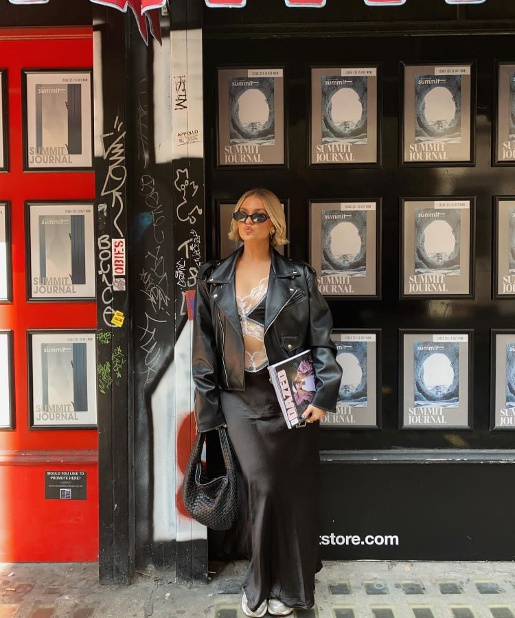 A foto mostra uma mulher em pé diante de uma parede cheia de cartazes com a inscrição "Summit Journal". Ela veste uma jaqueta de couro preta, saia longa e blouse transparente, carregando uma revista e usando óculos escuros. Há um contraste entre os elementos modernos e grafites na parede.