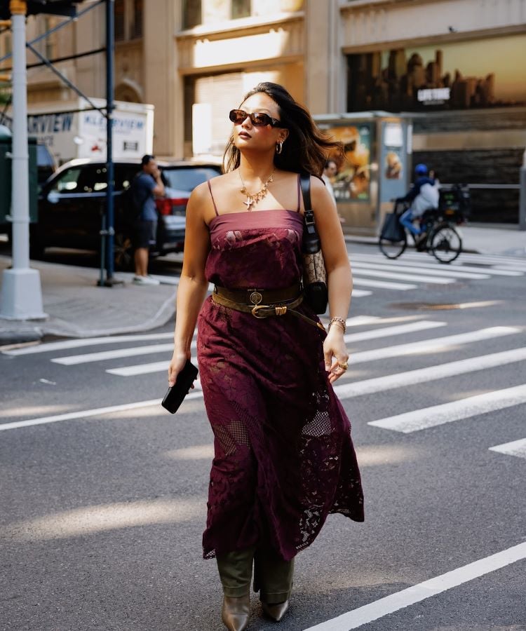 A imagem mostra uma mulher caminhando em uma rua da cidade. Ela usa um vestido longo bordô, cinto marrom, botas e óculos escuros. Seu cabelo está solto, e ela segura um celular na mão. A cena urbana ao fundo inclui carros e um ciclista, destacando um ambiente movimentado.