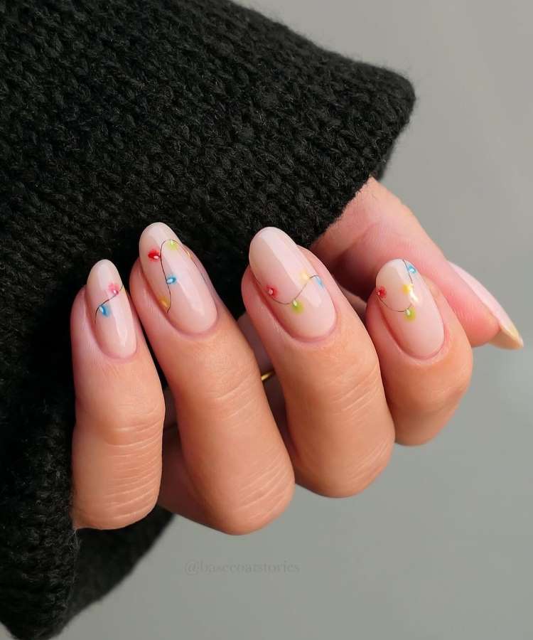 A imagem mostra uma mão com unhas pintadas de nude, decoradas com desenhos de luzes natalinas coloridas em vermelho, azul, verde e amarelo. As unhas são longas e elegantes. A mão está parcialmente coberta por um suéter preto de malha grossa.