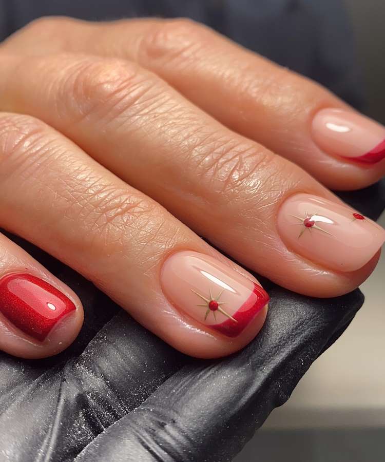 A imagem mostra uma mão com unhas decoradas em estilo minimalista. As unhas têm fundo nude e detalhes vermelhos, incluindo pontas francesas e pequenas estrelas centralizadas com pontos vermelhos. A mão é segurada por uma luva preta, destacando o design elegante.