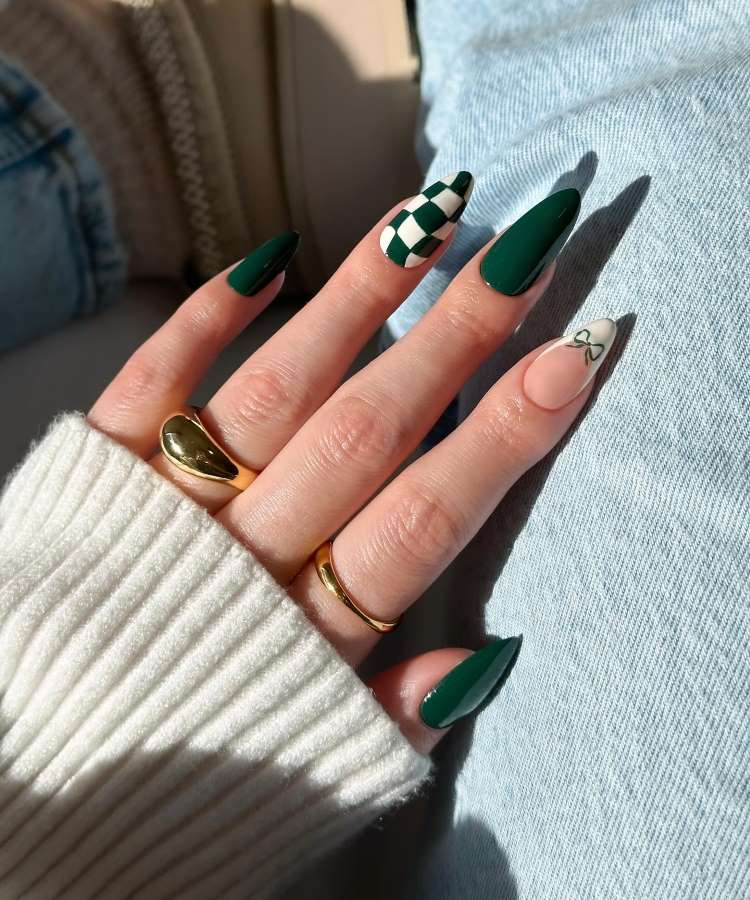 A imagem mostra uma mão com unhas longas e pontiagudas. Duas unhas são verdes sólidas, uma possui padrão quadriculado verde e branco, e outra tem desenho de laço sobre nude. Os dedos têm anéis dourados, e a mão veste um suéter creme.