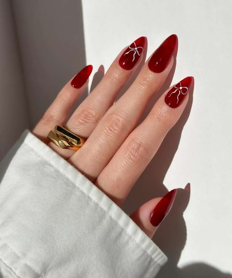 A imagem mostra uma mão com unhas longas, pintadas de vermelho brilhante. Duas unhas apresentam um desenho de laço branco. A mão usa um anel dourado moderno, e está parcialmente coberta por uma camisa branca. A luz destaca o brilho do esmalte.