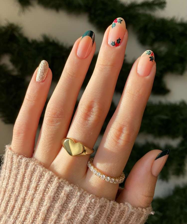 A imagem mostra uma mão com unhas elegantes e decoradas. Três unhas têm pontas pretas em estilo francês, outras apresentam flores vermelhas e verdes. Uma unha é coberta de glitter dourado. A mão usa anéis dourados, destacando-se sobre um suéter creme.