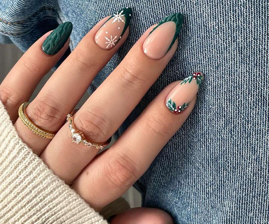 A imagem apresenta uma mão com unhas decoradas para o Natal. Duas unhas têm pontas verdes com flocos de neve brancos, uma com folhas verdes e frutos vermelhos. A pele é adornada por dois anéis dourados sobre um fundo de jeans.