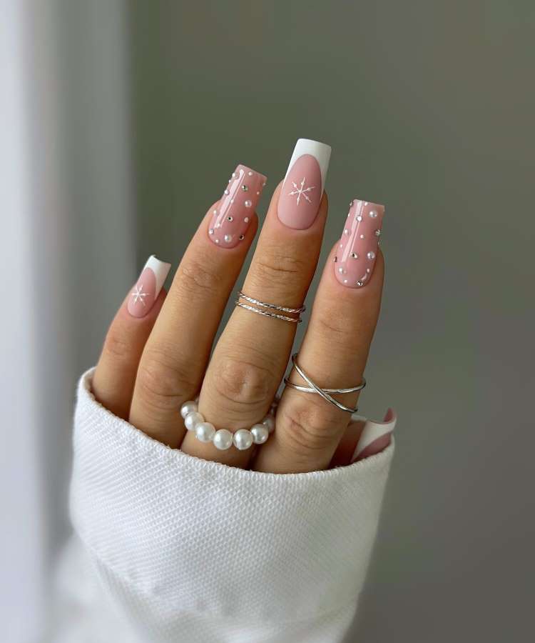 A imagem mostra uma mão com unhas decoradas em tons de rosa claro e branco, com detalhes de estrelas e pontos prateados. As unhas são longas e quadradas. A mão possui anéis prateados e de pérolas, enquanto veste um suéter branco.