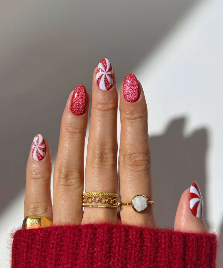 A imagem apresenta uma mão com unhas festivas. Duas unhas possuem listras vermelhas e brancas em espiral, lembrando doces natalinos. Outras são cobertas de glitter vermelho. A mão usa anéis dourados delicados e um suéter vermelho, em um cenário iluminado.