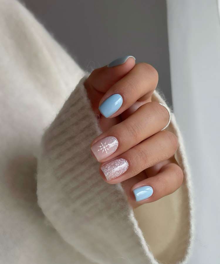 A imagem mostra uma mão com unhas pintadas em azul pastel e uma unha em rosa claro com glitter e floco de neve branco. O pulso está envolto em um suéter creme com textura suave. A mão exibe um delicado anel prateado.