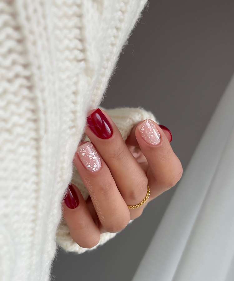 A imagem apresenta uma mão com unhas elegantes. Duas unhas são pintadas em vermelho profundo, enquanto as outras têm esmalte rosa claro com glitter, imitando um efeito de neve. A mão usa um anel dourado delicado e veste um suéter branco tricotado.