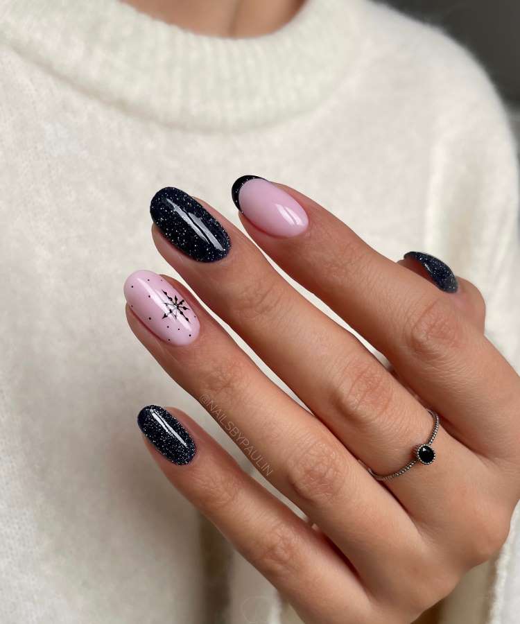 A imagem mostra uma mão com unhas decoradas em esmalte rosa claro e preto com glitter. Uma unha rosa tem um floco de neve desenhado. A mão usa um anel prateado com pedra preta e veste um suéter creme de lã.