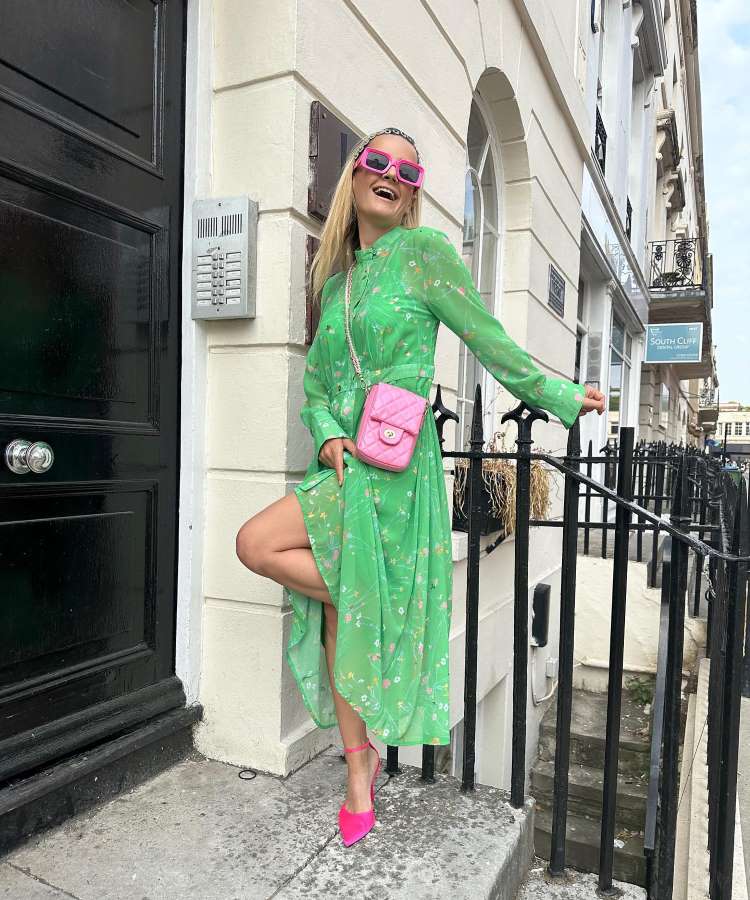 A imagem retrata uma mulher em estilo Wickedcore, com a tendência verde e rosa. Ela veste um vestido verde estampado com sapatos rosa vibrante e óculos escuros rosa. A bolsa rosa complementa o visual, posando alegremente em uma calçada urbana.