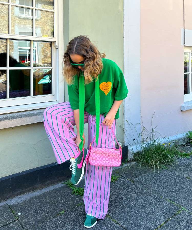 A imagem mostra uma pessoa com cabelo ondulado, óculos escuros e camisa verde com coração amarelo. Ela veste calças listradas rosa e verde, tênis verde e segura uma bolsa rosa. Está em frente a uma janela com plantas ao redor.