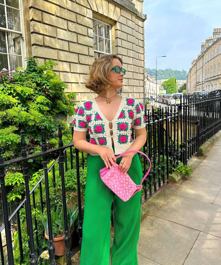 A imagem mostra uma mulher em um estilo Wickedcore, com tendência verde e rosa. Ela veste uma blusa de crochê com flores rosas, calças verdes vibrantes e segura uma bolsa rosa. Óculos verdes completam o look, em uma rua com arquitetura clássica.