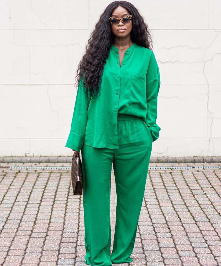 A imagem mostra uma mulher de cabelos longos e ondulados, usando óculos escuros com estampa de tartaruga. Ela veste um conjunto verde brilhante com camisa de botões e calças largas. Segura uma bolsa escura e está em pé sobre um piso pavimentado.