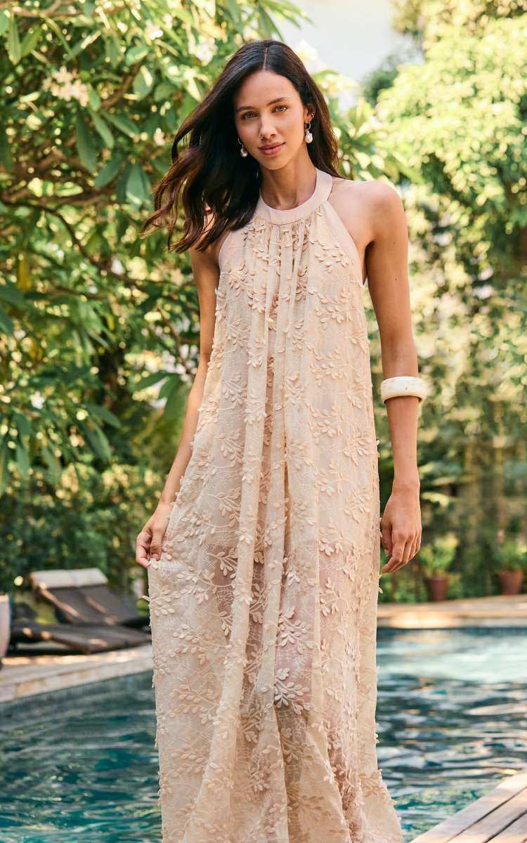 Esta peça da coleção Summer Breeza da Skazi apresenta um vestido longo bege com detalhes florais em renda. A modelo está ao lado de uma piscina, em um cenário tropical e elegante.