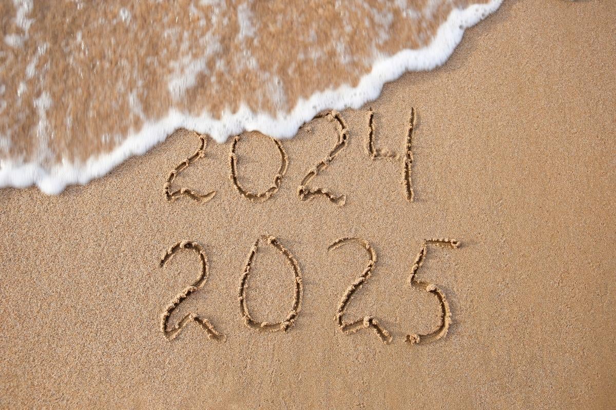 Escrito 2024 e 2025 na areia de praia com onda cobrindo parte de 2024. Retrospectiva 2024