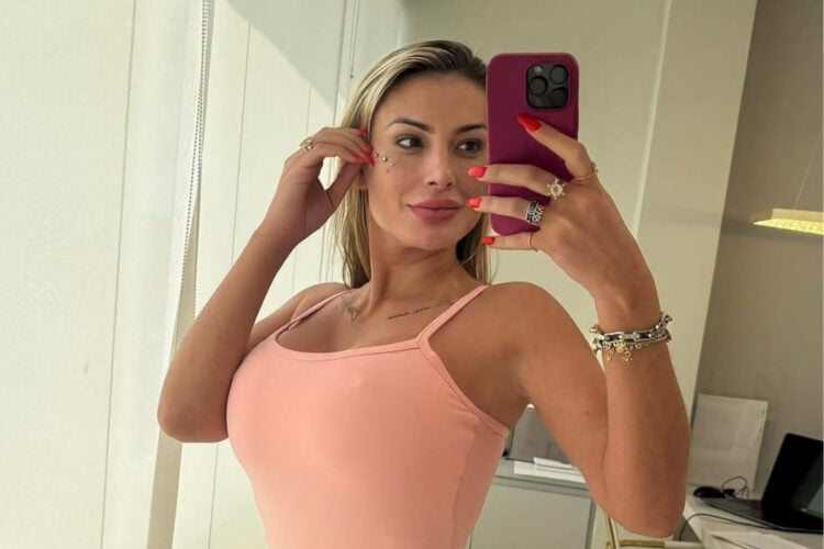 Selfie da influencer Andressa Urach em frente ao espelho usando macaquinho rosa e segurando celular com capinha roxa