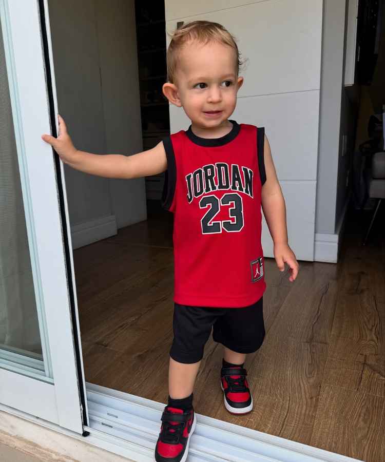 Um menino pequeno, vestindo uma camiseta vermelha do Jordan com o número 23, está na porta de vidro aberta. Ele está de pé sobre um piso de madeira, olhando curioso para o lado, usando tênis combinando.