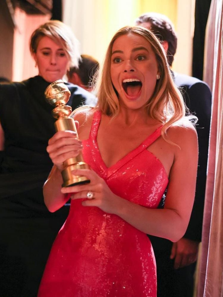 Foto de Margot Robie sorrindo, usando vestido rosa e segurando prêmio Globo de Ouro