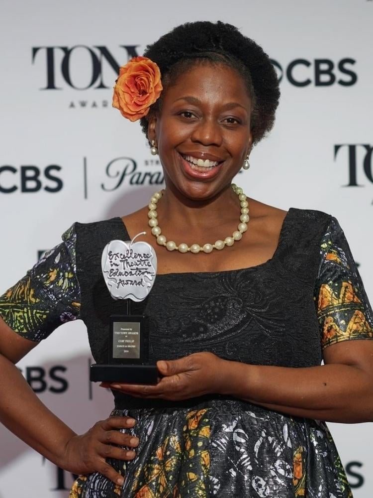 Mulher de pele negra usando vestido preto no colo e estampado nas mangas e saia, segurando um prêmio do The Tony Awards