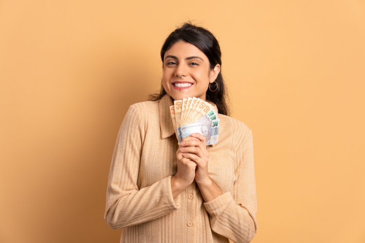 A imagem mostra uma mulher sorridente, de cabelos escuros e longos, segurando notas de euro em leque. Ela veste uma blusa bege, com um fundo laranja claro ao redor.