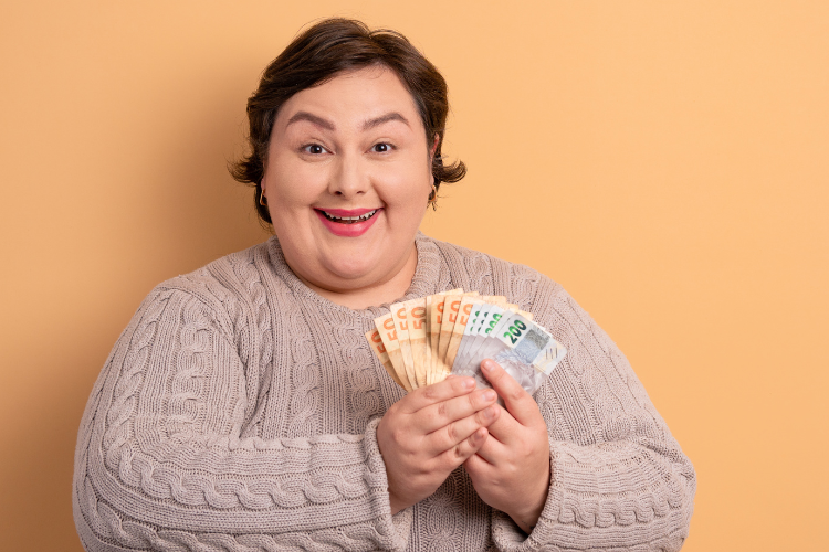 A imagem mostra uma pessoa sorridente segurando notas de euro. Ela veste um suéter bege, tem cabelo curto e expressão de alegria, com um fundo laranja claro ao redor.