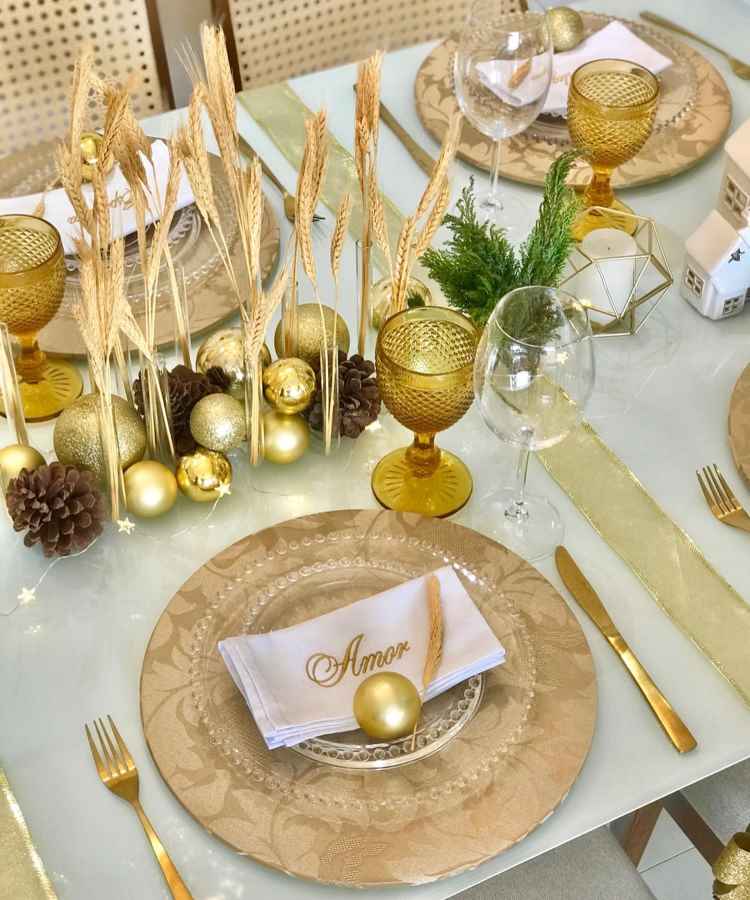 A imagem mostra uma mesa de Ano Novo elegantemente decorada. Louças douradas, taças âmbar, pinhas e bolas douradas decoram o centro. A mesa exibe guardanapos com "Amor" em dourado e detalhes naturais, criando um ambiente sofisticado e festivo para celebração.