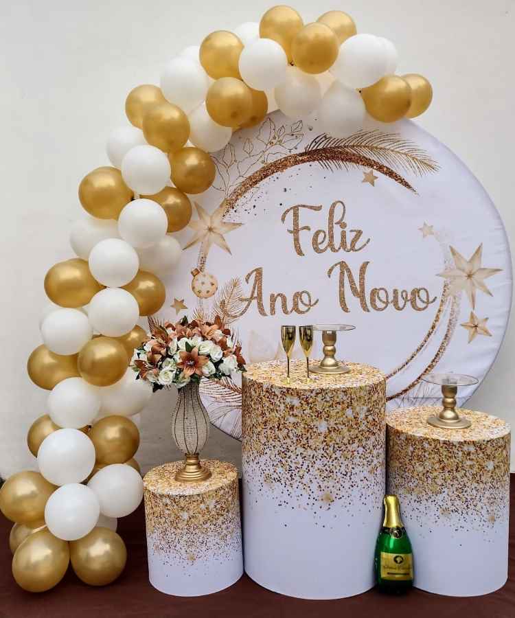 A imagem mostra uma decoração de Ano Novo com balões dourados e brancos formando um arco. Ao centro, um painel com "Feliz Ano Novo" em dourado. Três cilindros decorativos com glitter suportam taças douradas e uma garrafa de champanhe, completando o cenário festivo.