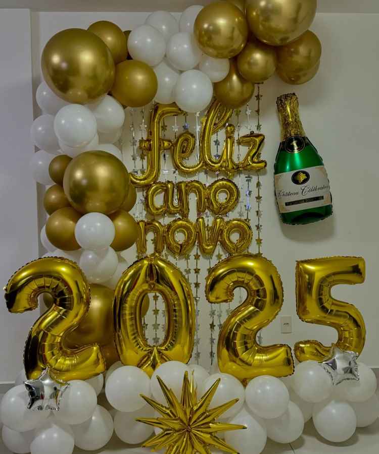 A imagem apresenta uma decoração de Ano Novo com balões dourados e brancos. As palavras "Feliz Ano Novo" estão destacadas, juntamente com o número "2025" em balões dourados. Há uma garrafa de champanhe inflável e estrelas decorativas complementando o cenário festivo.