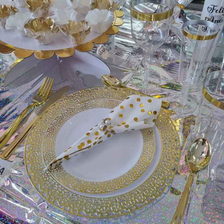 A imagem apresenta uma mesa sofisticada para Ano Novo com louças decoradas em dourado e branco. Talheres dourados acompanham pratos com detalhes intrincados. Um guardanapo branco com bolinhas douradas está dobrado centralmente, enquanto taças de vidro têm bordas douradas, criando um ambiente elegante e festivo.