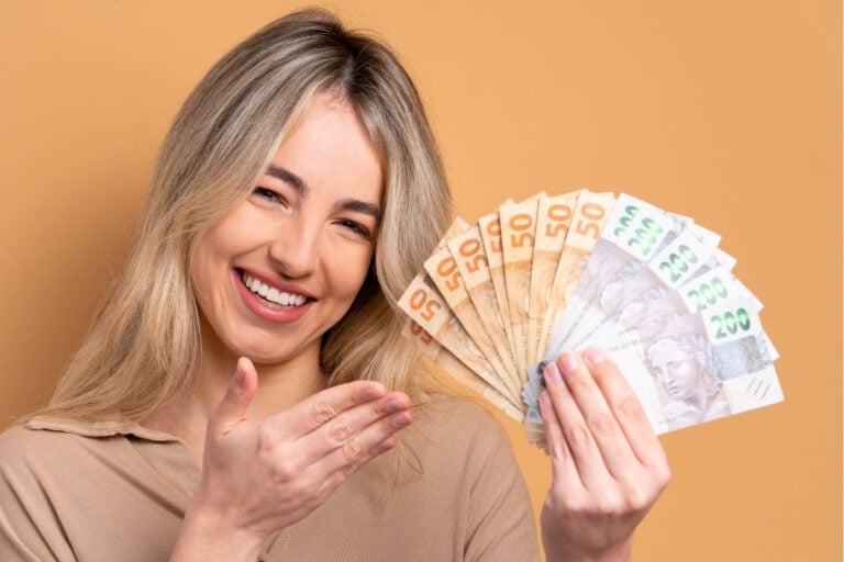 A imagem mostra uma mulher sorridente segurando notas de dinheiro em leque, aparentando felicidade. Ela está de frente, em um fundo bege, com cabelo loiro e expressão alegre.