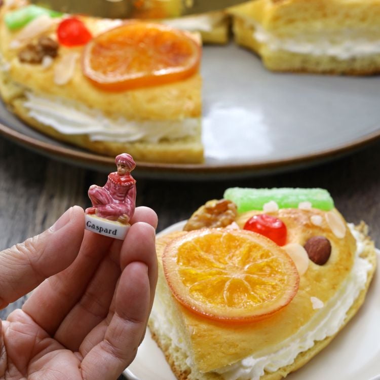 A imagem mostra uma fatia de bolo decorada com frutas cristalizadas, nozes e uma fatia de laranja. Em primeiro plano, uma mão segura uma pequena figura de porcelana, representando "Gaspar", um dos três reis magos. Ao fundo, mais fatias do bolo.