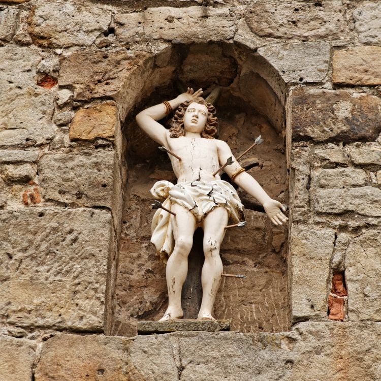 A imagem mostra uma escultura de São Sebastião, inserida em uma parede de pedra. Ele está amarrado e perfurado por flechas, simbolizando seu martírio. A estátua exibe uma expressão de serenidade, destacando sua resiliência e devoção religiosa.