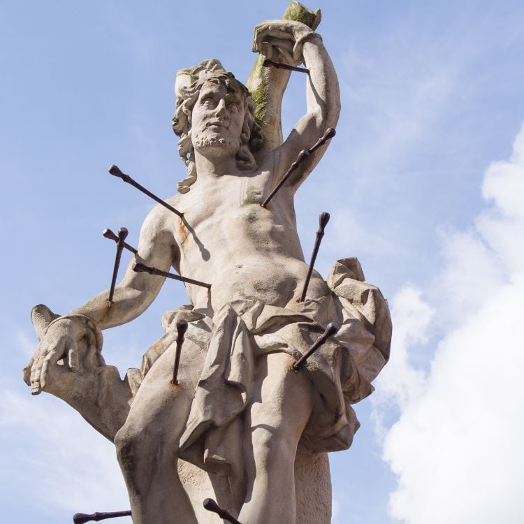 A imagem exibe uma estátua de São Sebastião, representado como mártir. Ele está amarrado, com várias flechas perfurando seu corpo. A escultura evidencia sua expressão serena e resiliência, destacando o céu ao fundo, simbolizando fé e sacrifício.