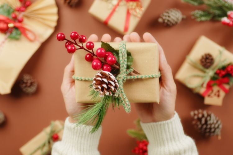 Mãos seguram um presente embrulhado em papel kraft com corda verde. O pacote é decorado com ramos de pinheiro, uma pinha e bagas vermelhas. Ao redor, outros presentes similares e pinhas sobre uma superfície marrom, criando um ambiente natalino acolhedor.