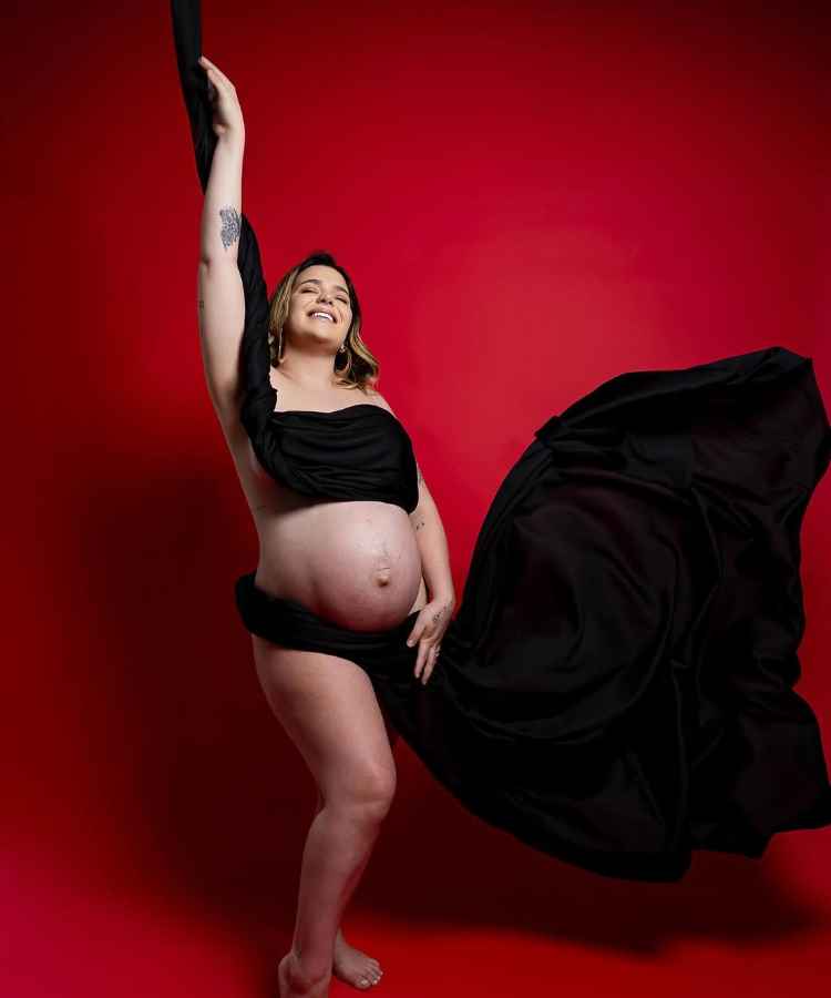 A imagem mostra uma mulher grávida em um estúdio com fundo vermelho. Ela está sorrindo, usando um tecido preto drapeado que envolve seu corpo e flui ao vento. A pose é confiante e artística, destacando a barriga e a expressão de felicidade - famosas que anunciaram gravidez