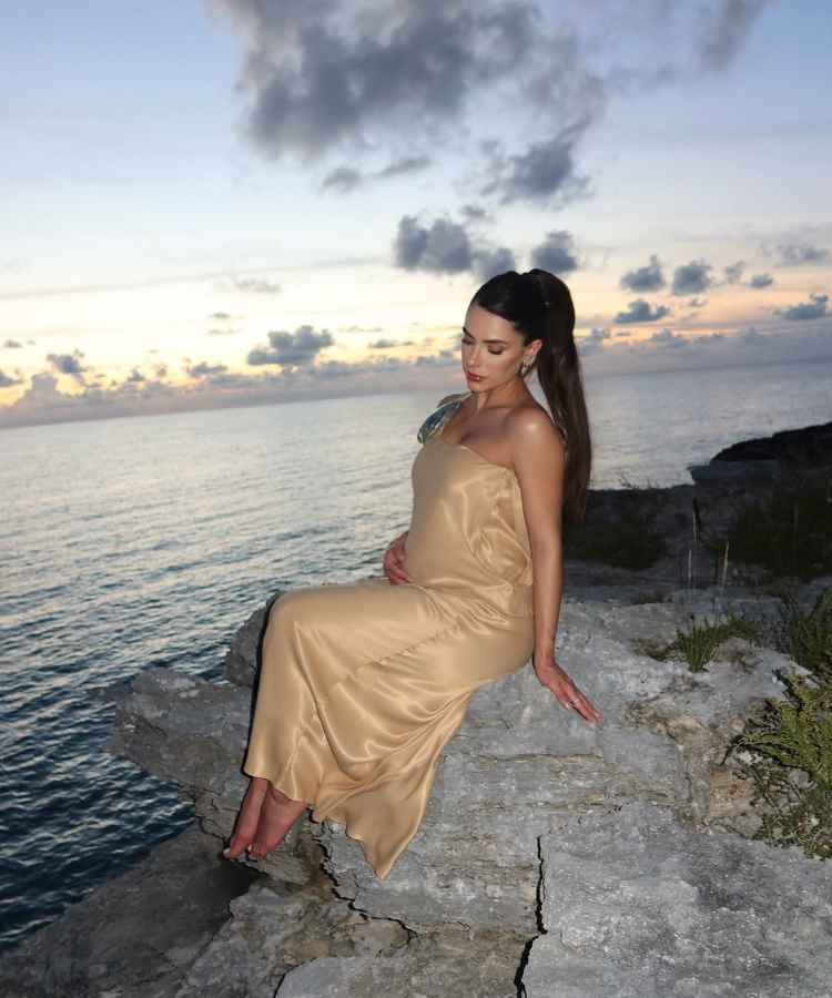 Uma mulher senta em uma rocha à beira do mar durante o pôr do sol. Ela usa um vestido longo dourado e olha suavemente para baixo. O mar e o céu, com nuvens ao entardecer, criam um cenário tranquilo e sereno.