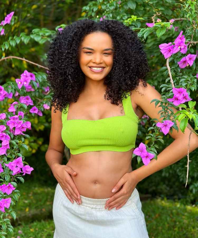 Uma mulher está sorrindo em meio a flores roxas e vegetação verde. Ela usa um top verde e saia branca, com as mãos repousando sobre a barriga. Seu cabelo é encaracolado e volumoso, e a cena transmite uma sensação de alegria e tranquilidade  - famosas que anunciaram gravidez