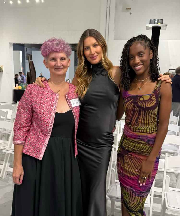 A imagem mostra três mulheres sorrindo em um evento social. A primeira usa casaco rosa e vestido preto, a segunda é Gisele Bundchen e veste vestido preto elegante, e a terceira um vestido colorido. Ao fundo, cadeiras brancas e uma parede clara.