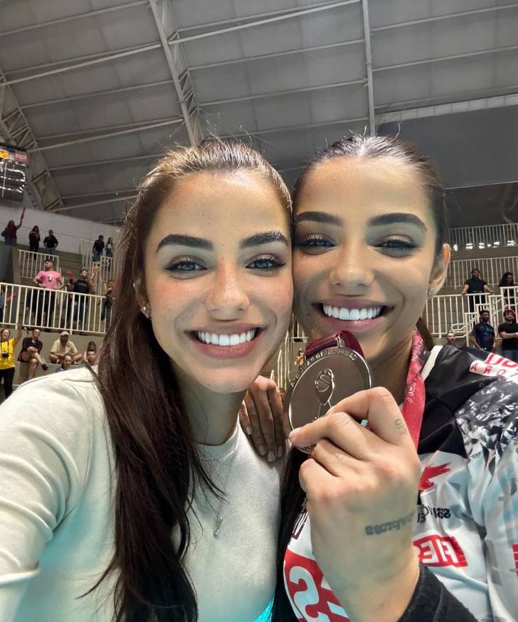 Duas mulheres sorridentes tiram uma selfie em um ambiente esportivo. Uma delas segura uma medalha, vestindo uniforme atlético. Ao fundo, pessoas assistem, cercadas por arquibancadas dentro de um ginásio.