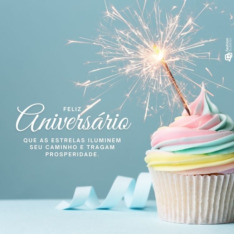 A imagem exibe um cupcake com cobertura de cores pastel, decorado com uma vela em forma de chama de artifício acesa. Ao lado, lê-se "Feliz Aniversário. Que as estrelas iluminem seu caminho e tragam prosperidade." O fundo é azul claro.