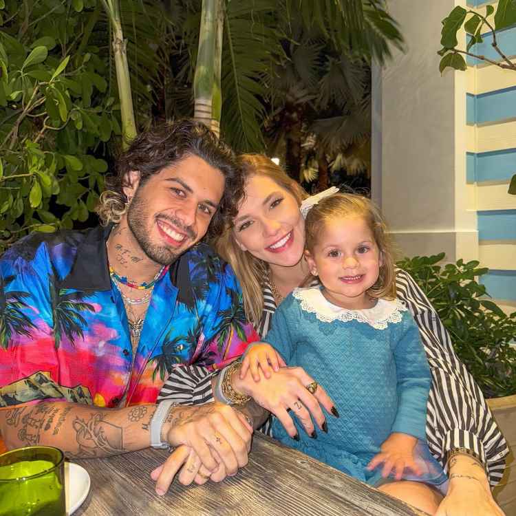 Um casal sorridente, com um homem de camisa colorida e mulher de blusa listrada, posa ao lado de uma menina com vestido azul e laço no cabelo. Eles estão sentados em uma mesa ao ar livre, cercados por plantas.