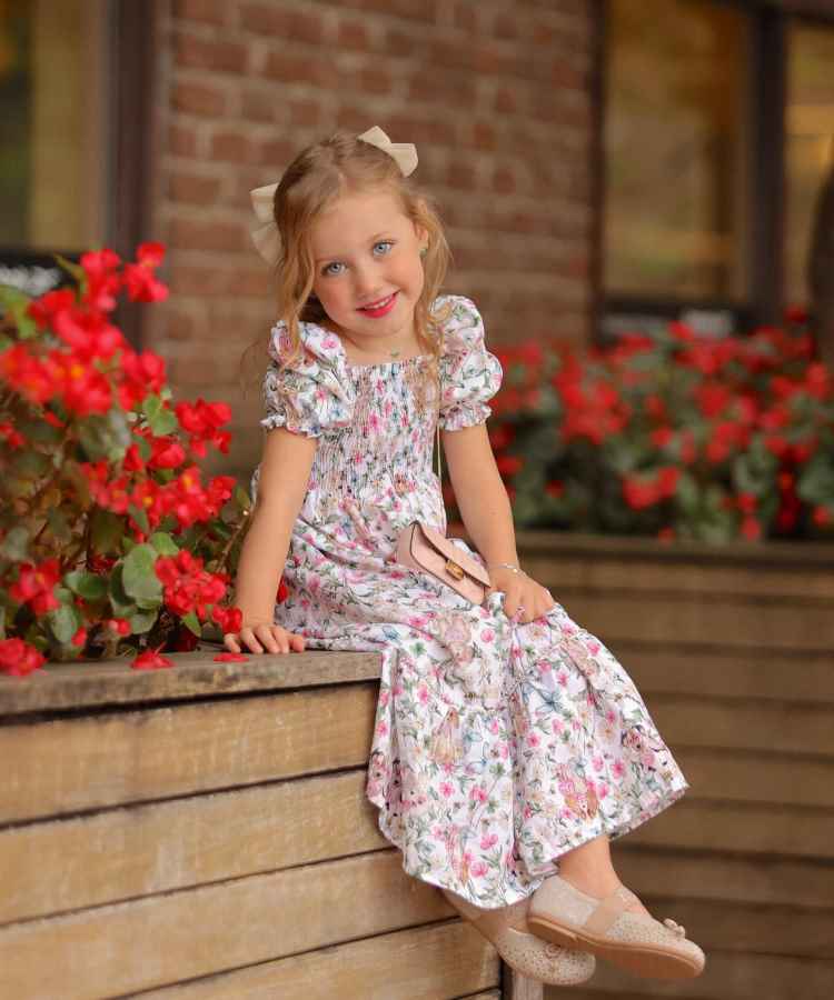 Uma menina loira, sorridente, veste um vestido floral e laço no cabelo, sentada em uma borda de madeira. Ao fundo, há flores vermelhas e uma parede de tijolos. Sua expressão é encantadora e cheia de alegria. filhos de famosos que fazem sucesso na internet