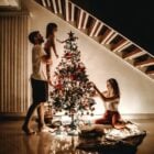 A imagem mostra uma família decorando uma árvore de Natal. Um homem segura uma criança, enquanto uma mulher ajusta as luzes.