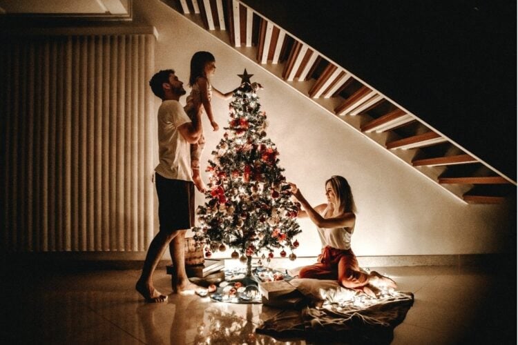 A imagem mostra uma família decorando uma árvore de Natal. Um homem segura uma criança, enquanto uma mulher ajusta as luzes.